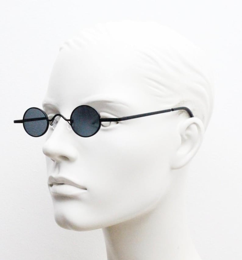 May include: Black round sunglasses with thin black frames. The lenses are dark, and the glasses have a minimalist design. The sunglasses are displayed on a white mannequin head.