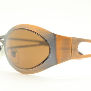 Op de afbeelding: Ovale zonnebril met een bronskleurig montuur en bruine glazen. Het montuur heeft een uniek ontwerp met een donkergrijze rand rond de glazen en een uitgesneden detail op de veren. Een stijlvol accessoire.