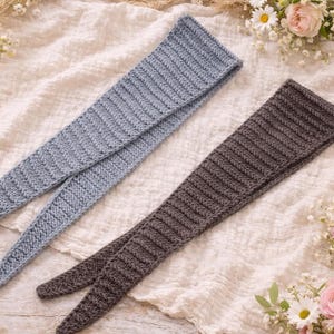May include: Two knitted scarves. One is pale blue, the other is brown. Both have a ribbed texture and a pointed end. The scarves are displayed on a cream fabric, with flowers and knitting needles in the background.