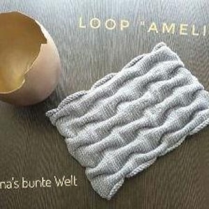 May include: Gray knitted loop scarf with a textured pattern. The scarf is laid flat on a wooden surface. The text "LOOP "AMELIE" is visible in the upper right corner of the image.