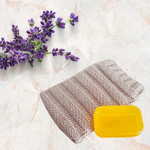 May include: A sprig of purple lavender rests beside a beige knitted washcloth and a yellow bar of soap. The soap has text on it. The items are arranged on a marble surface.