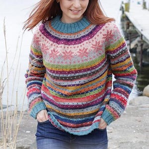 Könnte beinhalten: Ein farbenfroher Strickpullover mit Rundhalsausschnitt und langen Ärmeln. Der Pullover hat ein Muster aus horizontalen Streifen in verschiedenen Farben, darunter Blau, Rosa, Grün und Lila. Der Text "DROPS DESIGN" ist unten im Bild zu sehen.
