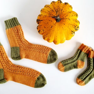 May include: Three pairs of knitted socks in a mustard yellow and forest green colour scheme. The socks are arranged on a white surface with a yellow squash in the background.