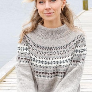 Könnte beinhalten: Ein hellgrauer Pullover mit hohem Kragen und einem gemusterten Passe. Die Passe hat ein geometrisches Design in Weiß, Braun und Blau. Das Bild enthält auch den Text "DROPS DESIGN" und die Website-Adresse.