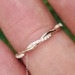 May include: A delicate gold band ring with a twisted design and small clear stones set along the band. The ring is on a finger.