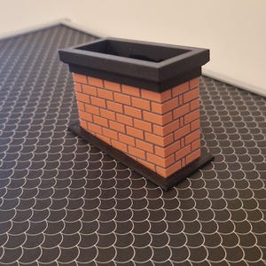 Puede incluir: Una pequeña chimenea de ladrillo con la parte superior e inferior negras. La chimenea está hecha de ladrillos naranjas y se asienta sobre una superficie oscura, gris y con diseño de escamas. Es un modelo o un objeto decorativo.