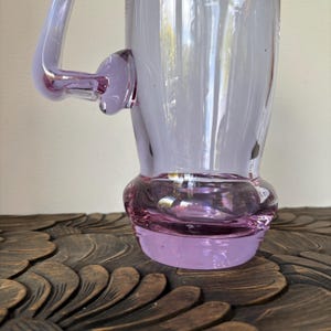 May include: A tall, lavender-colored glass mug with a handle. The mug has a bulbous base and a flared rim. The handle is curved and attached to the side of the mug. The glass is transparent, allowing light to pass through. The mug is sitting on a wooden surface.