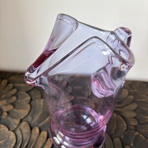 May include: A clear, lavender-colored glass vase with a unique, asymmetrical shape. The vase has a flared rim and a textured, ridged base. The glass is transparent, allowing light to pass through, and the color is a soft, delicate purple.
