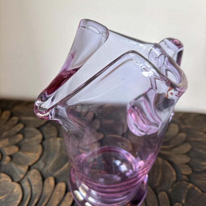 May include: A clear, lavender-colored glass vase with a unique, asymmetrical shape. The vase has a flared rim and a textured, ridged base. The glass is transparent, allowing light to pass through, and the color is a soft, delicate purple.
