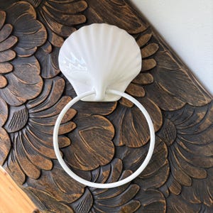 May include: A white ceramic seashell-shaped towel ring holder. The holder features a circular ring for towels and is mounted on a dark brown, carved wooden surface with a floral pattern. The design evokes a coastal or nautical theme.