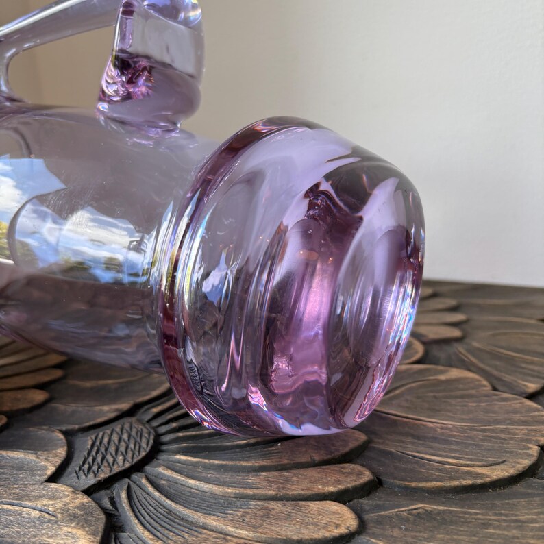 May include: A close-up of a lavender-colored glass pitcher with a handle. The pitcher has a bulbous base with a swirled design. The glass is translucent, allowing light to pass through. The pitcher sits on a dark, carved wooden surface.