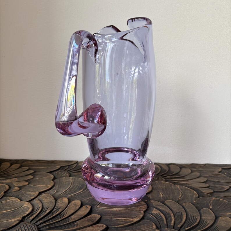 May include: A lavender glass vase with a unique handle and a bulbous base. The vase has a flared top and a decorative, rounded base. The handle curves out and around the side of the vase. The vase is sitting on a dark wooden surface.