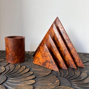 May include: A wooden desk organizer set featuring a cylindrical pen holder and a triangular letter holder. Both items display a warm, reddish-brown burl wood grain pattern. The set is displayed on a dark, carved wooden surface.