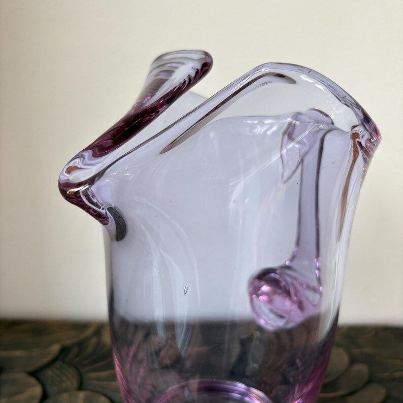 May include: A handcrafted, clear glass vase with a unique, ruffled rim and a delicate, lavender hue. The vase features a rounded, bulbous base and a gracefully curved handle-like detail. The glass has a smooth, polished finish, reflecting light and adding a touch of elegance.