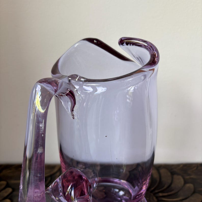 May include: A clear, lavender-colored glass pitcher with a curved handle and a unique, asymmetrical rim. The pitcher's design features a blend of light and dark purple hues, creating a visually appealing aesthetic. The pitcher is likely used for serving beverages.