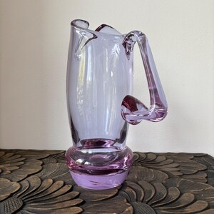 May include: A lavender-colored glass vase with a unique handle design. The vase has a bulbous base and a tall, slender body. The handle extends from the top and curves down to meet the body. The vase is made of transparent glass and is likely used for decorative purposes.