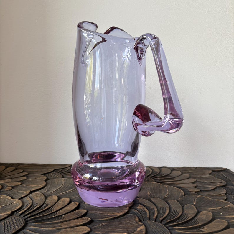 May include: A lavender-colored glass vase with a unique handle design. The vase has a bulbous base and a tall, slender body. The handle extends from the top and curves down to meet the body. The vase is made of transparent glass and is likely used for decorative purposes.