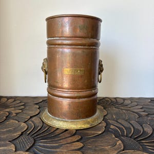 May include: A cylindrical copper ice bucket with a brass base and two brass lion head handles featuring hanging rings. A rectangular brass plaque on the front is engraved with the word WHISKY. The metal surface shows a weathered patina.