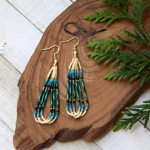 May include: Handmade beaded earrings with a gold hook. The earrings feature a cream-colored top section, transitioning to teal and dark green beads. The design is finished with a cream-colored bottom section. The earrings are displayed on a wooden surface with green foliage.