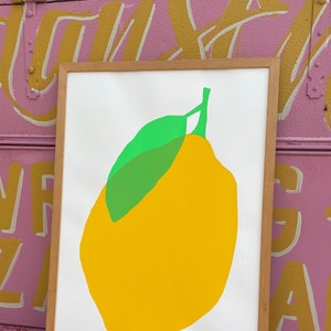 Puede incluir: Una impresión enmarcada de un limón amarillo con una hoja verde sobre un fondo blanco. La impresión está firmada "24/35".