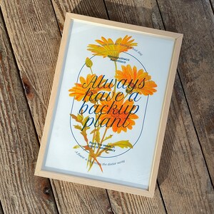 Op de afbeelding: Een ingelijst print met een gele bloemenillustratie en de tekst "Always have a backup plant. Plant the seeds in case of emergency. A flower a day keeps the doctor away."