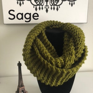 May include: Sage green knitted infinity scarf. The scarf is made of a soft, chunky yarn and has a textured stitch pattern.