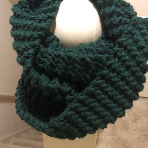 May include: A dark green knitted infinity scarf. The scarf is made with a chunky yarn and has a textured stitch pattern.