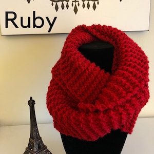 May include: A red knitted infinity scarf. The scarf is folded in half and displayed on a black mannequin stand. The word "Ruby" is written in black text on a white background.