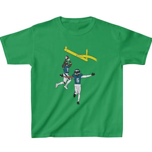 May include: Green t-shirt with a graphic of two football players in action. The players are wearing blue and white uniforms and are jumping for the ball. The graphic is set against a yellow goal post.