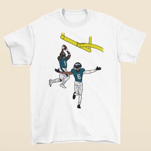 以下が含まれることがあります： 緑と白のユニフォームを着て、1と6の番号をつけた2人のフットボール選手がアクションを起こしているグラフィックが描かれた白いTシャツ。グラフィックは黄色のゴールポストを背景にしている。