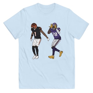 May include: A light blue t-shirt with a cartoon illustration of two football players. One player is wearing a purple and yellow uniform with the number 18, and the other player is wearing a black and orange uniform with the number 1.