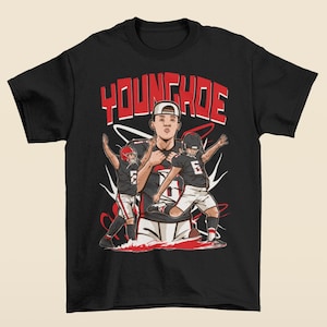 Puede incluir: Camiseta negra con un diseño gráfico que presenta a un jugador de fútbol americano y el nombre "YOUNGHOE" en rojo. El diseño incluye ilustraciones de jugadores de fútbol americano en acción, con detalles en rojo y blanco.