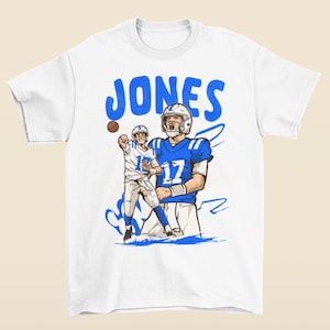 May include: White t-shirt featuring a blue graphic of a football player throwing a ball, with the name "JONES" in blue above the image. The player is wearing a blue and white uniform with the number 17.