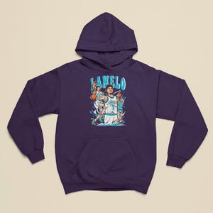 以下が含まれることがあります： 紫色のフード付きスウェットシャツ。バスケットボール選手のグラフィックと「LAMELO」という名前が水色で描かれています。グラフィックはバスケットボールをする選手を描いています。スウェットシャツにはフロントポケットが付いています。