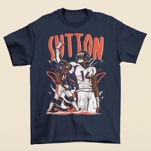 May include: Navy blue t-shirt featuring a graphic of a football player in a white and blue uniform, with the name "SUTTON" in orange above. The player is raising his arm. The number "14" is visible on the jersey.
