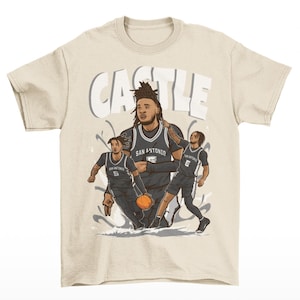 May include: Beige t-shirt featuring a graphic of a basketball player in a San Antonio Spurs jersey, holding a basketball. The word "CASTLE" is printed above the player. The design includes two additional players in action poses.