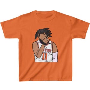 May include: Orange short-sleeved t-shirt with a cartoon illustration of a basketball player wearing a white jersey with the text "NYORK" and the number "11" on it. The player is covering their face with their hand.