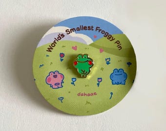 May include: A green frog pin with red accents and gold trim is centered on a circular card. The card features a green field with cartoon frogs, flowers, and the text "World's Smallest Froggy Pin" and "dahaae". The background is light blue.