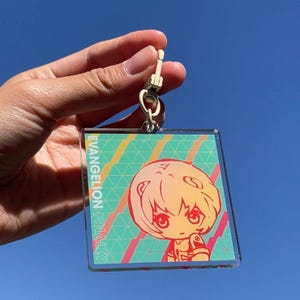 Peut inclure: Porte-clés carré avec un personnage de dessin animé aux cheveux orange et jaunes, aux yeux rouges et aux vêtements orange. Le porte-clés a un fermoir doré et le texte "EVANGELION FINALLY". Le fond est un ciel bleu.