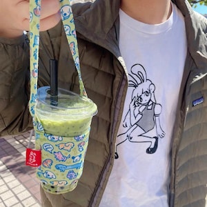 Puede incluir: Un vaso de plástico transparente lleno de una bebida verde, sostenido en un soporte de tela verde claro con una correa. El soporte está decorado con ilustraciones de perros coloridos. Una pajita negra está insertada en el vaso.