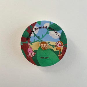 Peut inclure: Épingle ronde avec trois singes de dessin animé sur un fond vert et bleu. L'épingle porte l'inscription "World's Smallest Monkey Pin" et le nom de la marque "dahaae". Les singes sont roses, oranges et dorés.