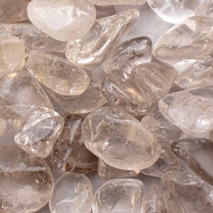 May include: A collection of clear quartz tumbled stones. The stones are smooth and polished, with a variety of shapes and sizes. They are a light brown color, with some stones having a more translucent appearance.