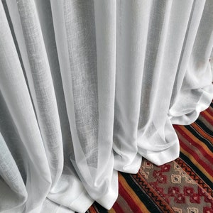 Puede incluir: Cortinas blancas transparentes con un tejido texturizado, drapeadas en suaves pliegues. Las cortinas cubren parcialmente una alfombra colorida con un patrón geométrico, creando un contraste de texturas y colores.