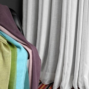 Puede incluir: Una pila de paneles de tela transparente de colores drapeados sobre una silla negra frente a una cortina blanca transparente. Los paneles de tela son en tonos de morado, rosa, verde, azul y turquesa.