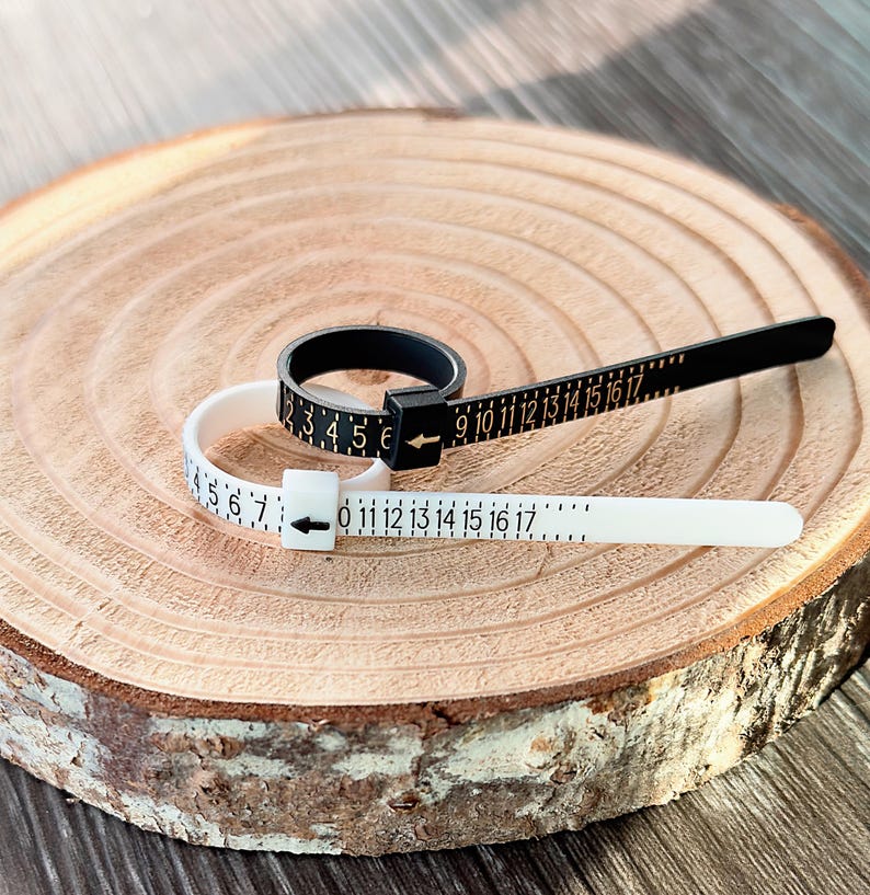 May include: Two ring sizers, one black and one white, with black numerals and arrows, are displayed on a wooden surface. The black sizer has numbers from 2 to 17, while the white sizer has numbers from 0 to 17. These tools are used to measure ring sizes.