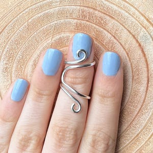 May include: A silver-colored, adjustable finger ring with a swirling design. The ring wraps around the finger with a decorative spiral at the top. The background is a light brown, wooden surface.