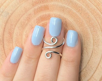 May include: A silver-colored, adjustable ring with a swirling design. The ring is worn on a finger with light blue nail polish. The background is a light brown, wooden surface.