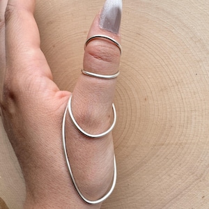 May include: A silver thumb ring with a unique design. The ring features two bands on the thumb and a larger, elongated loop that curves around the side of the thumb. The jewelry is displayed on a wooden surface.
