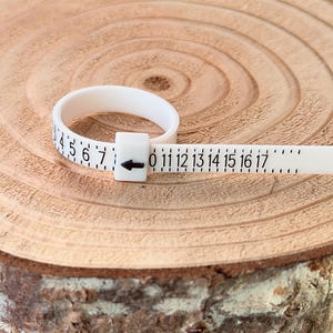 May include: A white ring sizer tool with black numbers and markings. The tool is used to measure ring sizes, with numbers from 0 to 17. The ring sizer is made of a flexible material and is placed on a wooden surface.