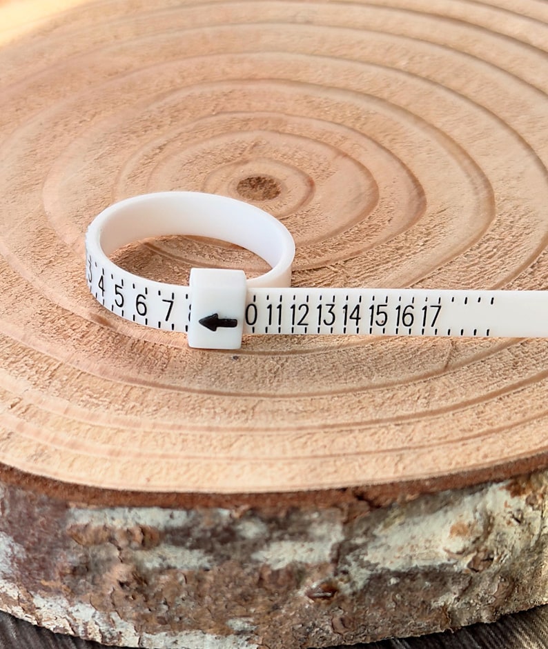 May include: A white ring sizer tool with black numbers and markings. The tool is used to measure ring sizes, with numbers from 0 to 17. The ring sizer is made of a flexible material and is placed on a wooden surface.